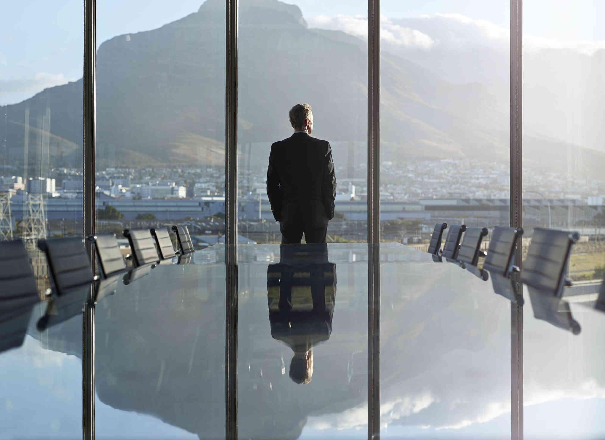 A man in a suit stands in a conference room with large windows as he gazes out of the window deep in thought with his hands in his pocckets.